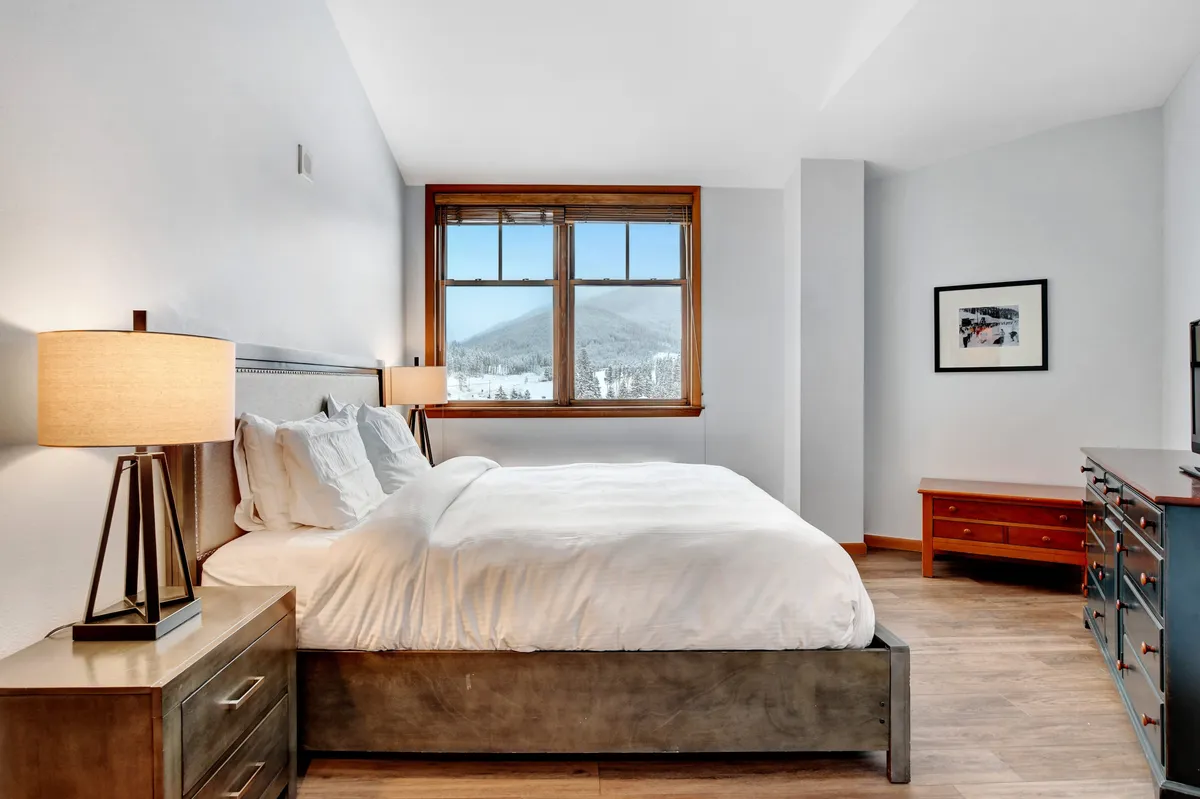 A serene bedroom with white linens, large windows offering a mountain view, lamps on bedside tables, and a wooden dresser. Natural light fills the space, creating a tranquil and cozy retreat.
