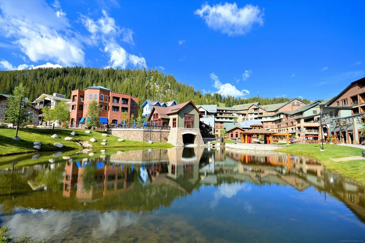 A cheerful lakeside scene featuring colorful buildings, reflective water, green landscape, and a majestic mountain backdrop. Guests are seen walking along the water, exploring local shops and restaurants, and enjoying the natural surroundings, suggesting a vibrant community atmosphere.