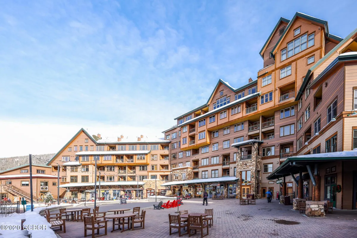 The exterior of a large, multi-story lodge with outdoor seating and vibrant red lounge chairs. People are walking and socializing, with retail shops and a welcoming lodge facade suggesting easy access to amenities and a lively community atmosphere.
