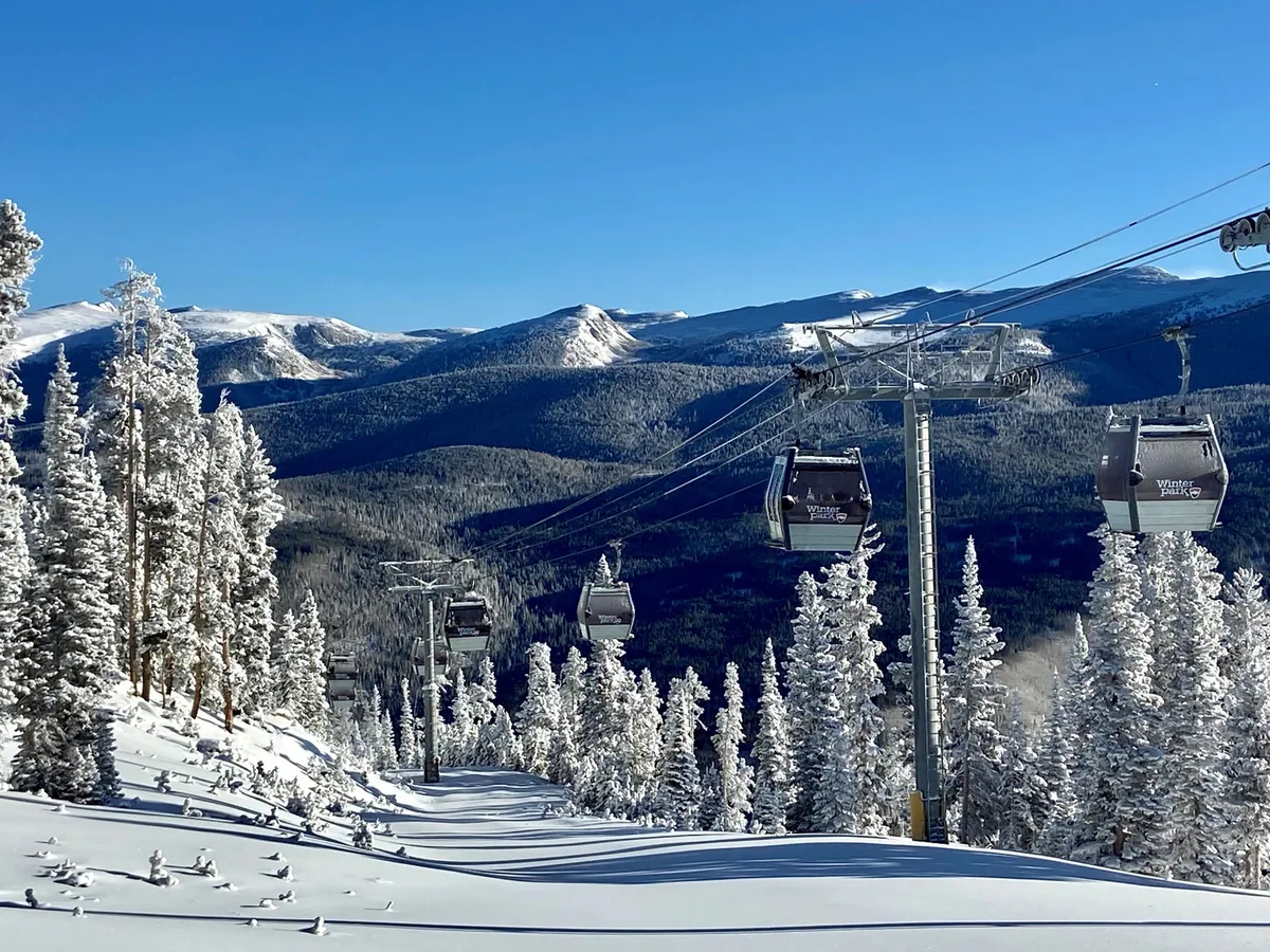 A snowy landscape featuring a ski lift ascending through tall, snow-covered trees with a majestic mountain range in the background. Skiers and snowboarders can be seen gliding down the slopes, highlighting the thrill and accessibility of the ski-in/ski-out experience.