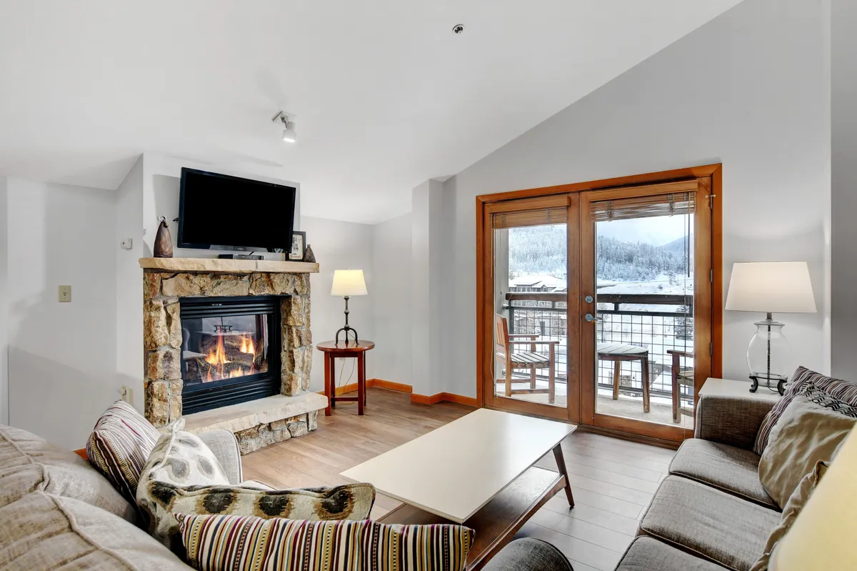 The image shows a stylish and inviting living room with a cozy fireplace as the centerpiece, surrounded by plush seating. Large windows flood the space with natural light and frame breathtaking snowy mountain views, while a coffee table invites gatherings and relaxing moments.