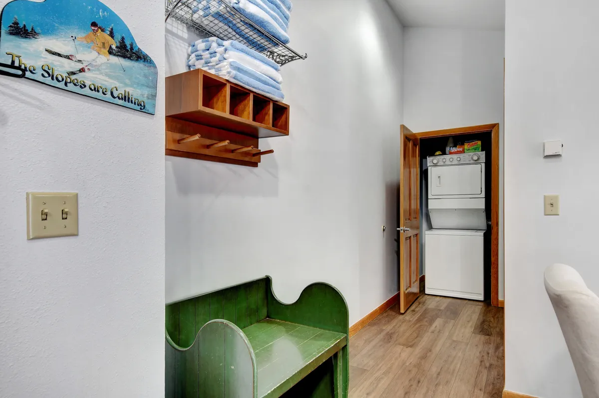 A mudroom or entryway featuring a green bench, laundry area, neatly stacked towels, and a sign reading 'The Slopes are Calling.' The space feels both practical and inviting, suggesting a convenient spot for guests to store gear and transition from outdoor to indoor activities.