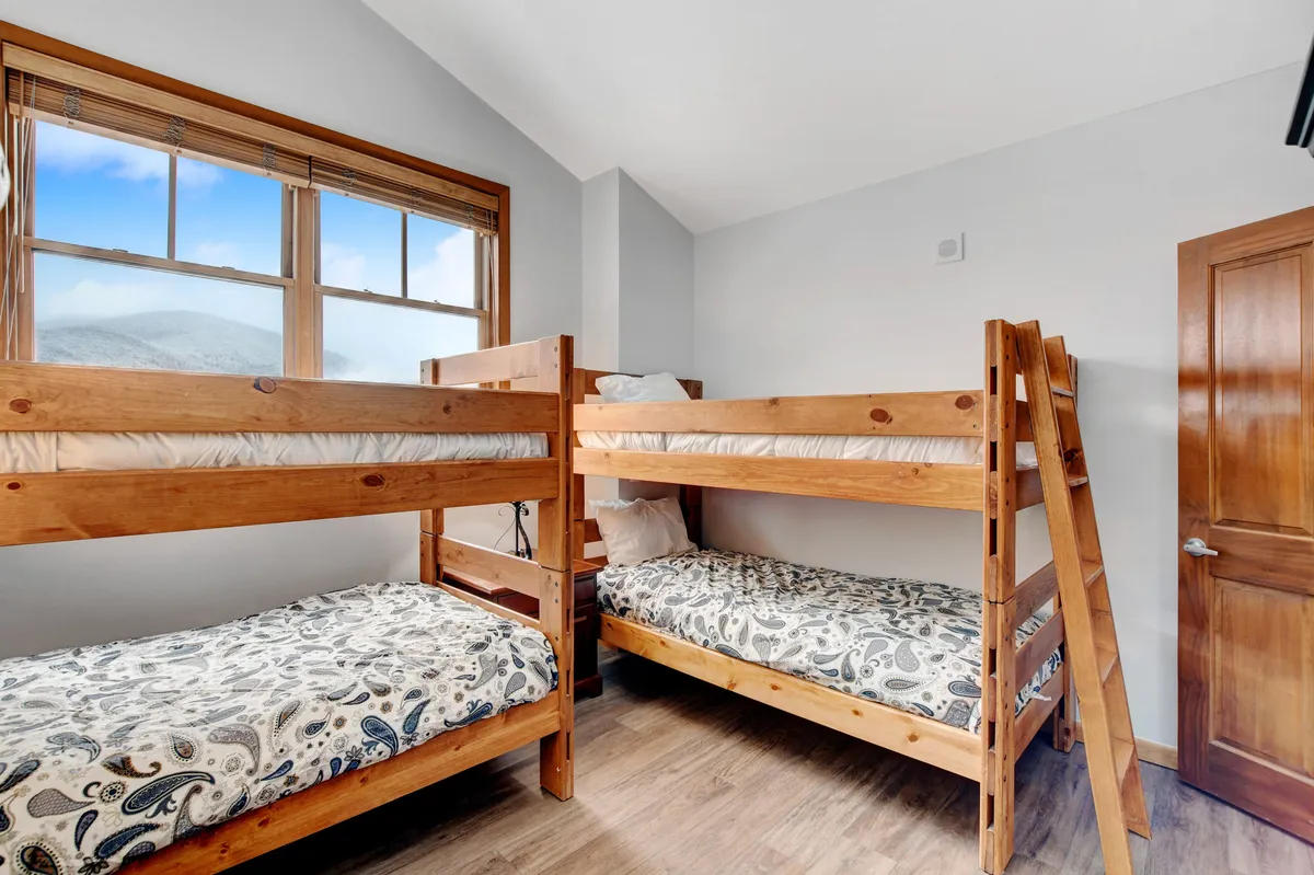 The photo features a cozy bunkroom with wooden bunk beds, a window offering natural light, and sturdy wooden furniture. The setting is ideal for children or friends, inviting activities like playing games, reading, or sharing stories.