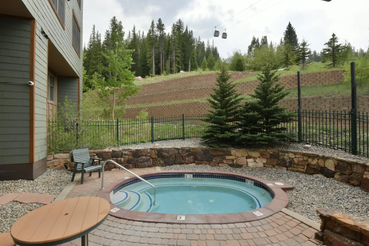 An inviting outdoor hot tub set against lush greenery, with a secure fenced area and dramatic mountain backdrop. The gondola in the sky hints at year-round mountain adventure, and the scene is perfect for relaxation or socializing.