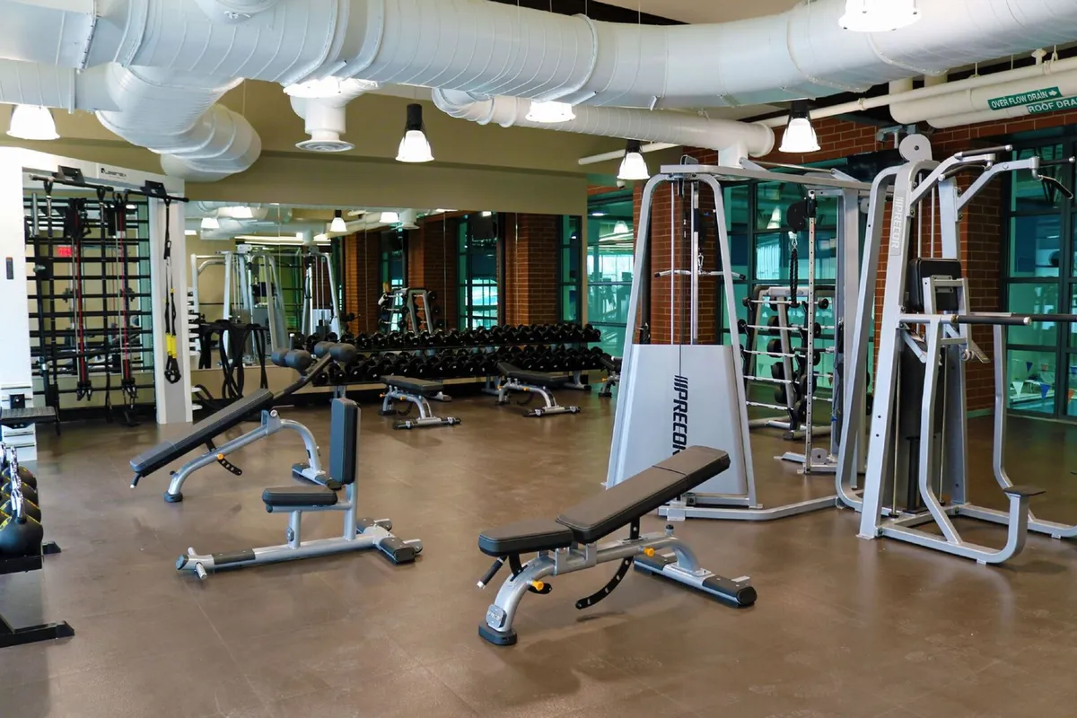 A modern, well-equipped gym with a weightlifting bench, dumbbell racks, exercise machines, and large mirrors. The space is designed for both individual and group fitness, fostering a motivating and energetic environment.