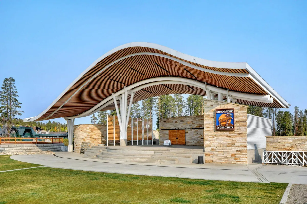 The image features an iconic curved roof structure and stone facade, with signage indicating an event center. It shows a space designed for outdoor concerts, community events, and family gatherings, bustling with people and activity.