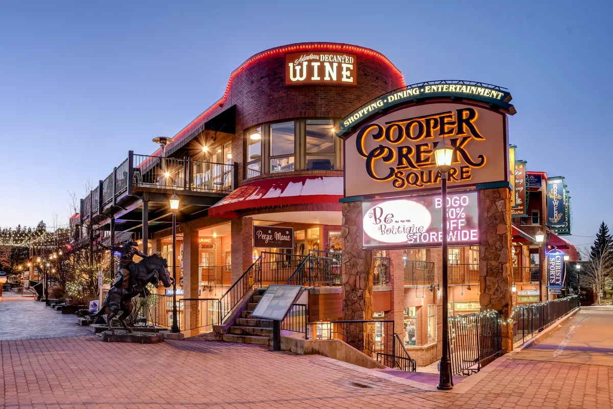 This exterior image showcases the lively Cooper Creek Square, featuring festive string lights, a wine shop sign, outdoor seating, and a prominent horse statue. The setting is bustling and inviting, hinting at the variety of shopping, dining, and entertainment experiences available in the heart of Winter Park Village.
