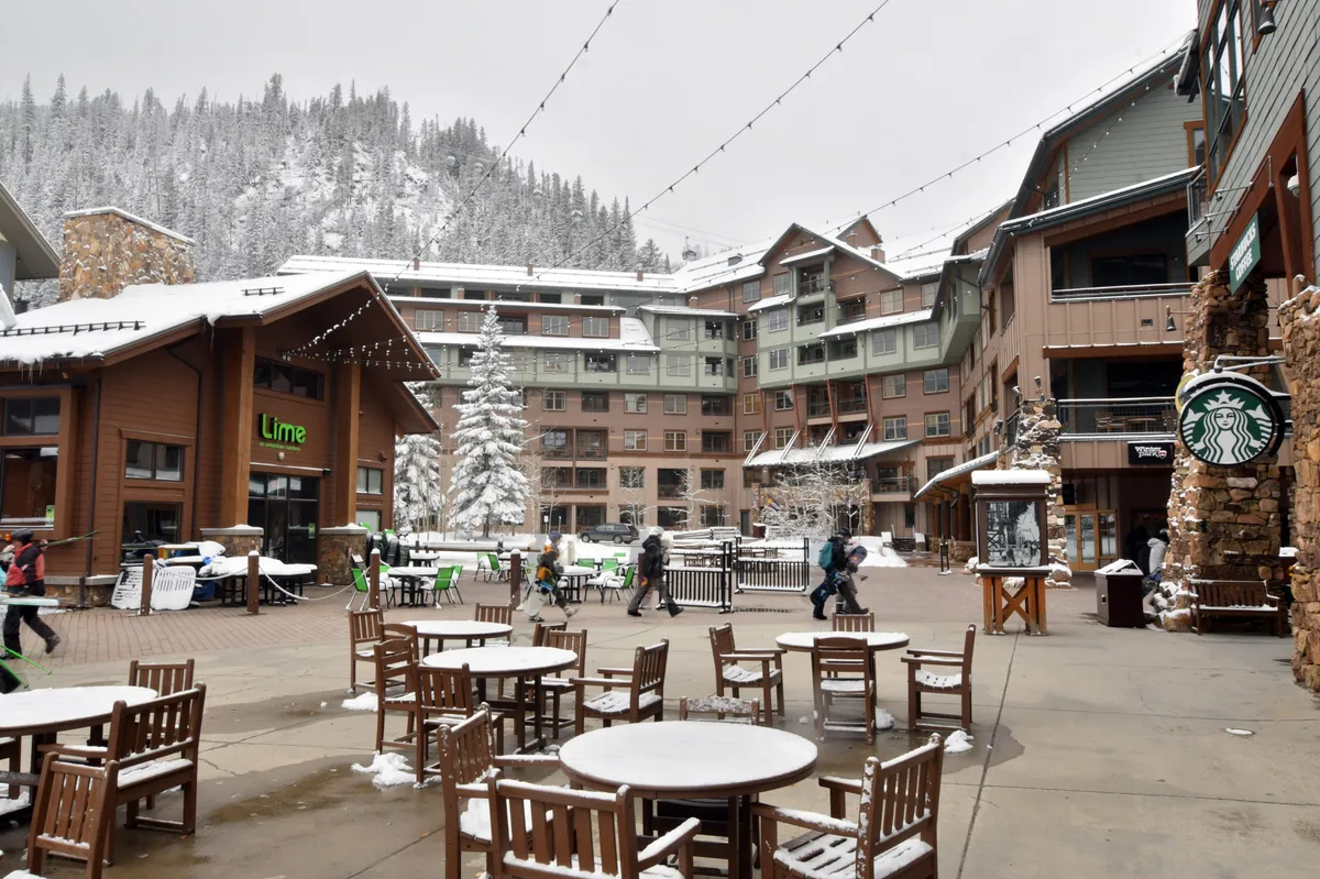 A cozy, festive exterior scene with snow-covered trees, outdoor seating areas at Lime restaurant and Starbucks, and people enjoying warm beverages. The atmosphere is inviting, with gatherings around outdoor heaters and a palpable sense of winter celebration.