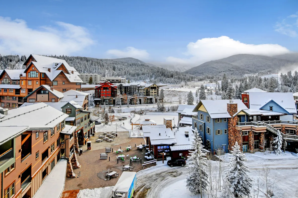 Colorful, snow-covered buildings with outdoor seating and a vibrant mountain village atmosphere. Snowy mountains provide a dramatic backdrop, and the scene is lively with signs of winter sports and après-ski activities.