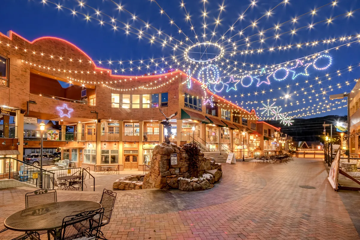 This exterior photo focuses on decorative string lights above a stone fountain and multiple storefronts, evoking a sense of community and festivity. People can be seen shopping, dining, and socializing in this bright, open-air village setting.