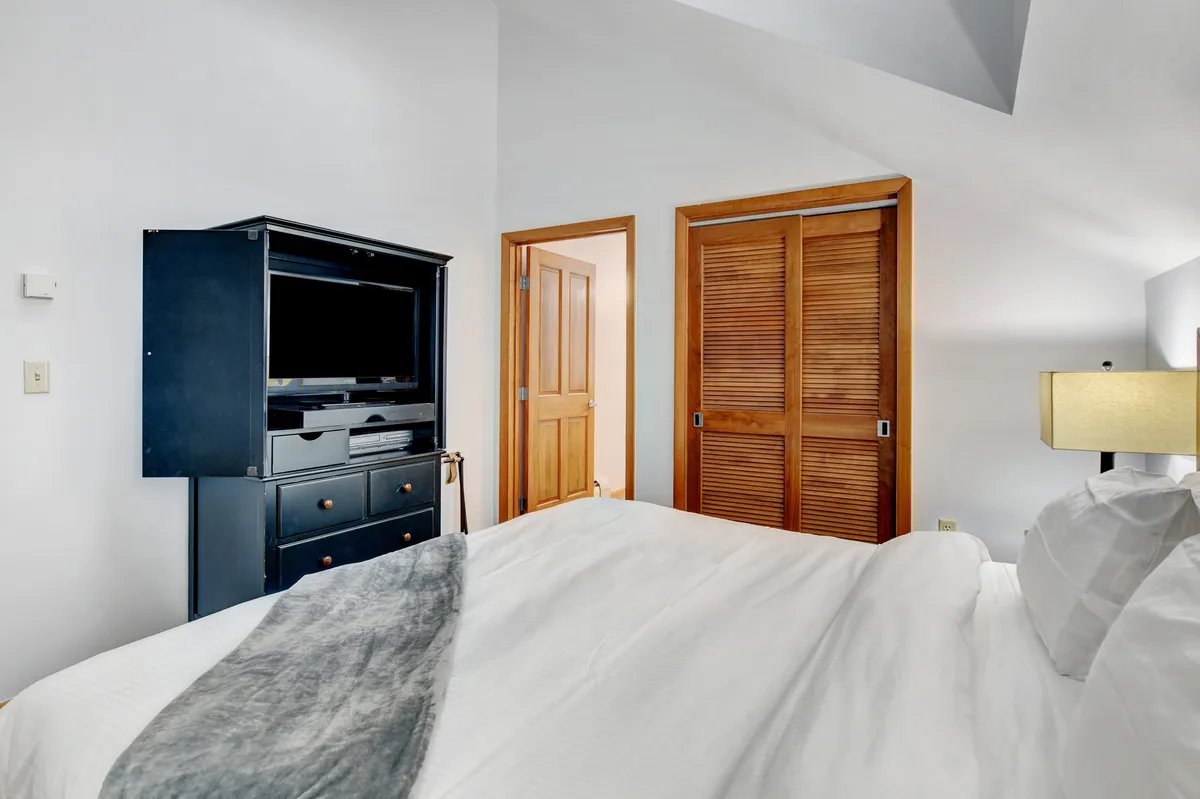 A welcoming bedroom featuring a comfortable bed, a television unit, a bedside lamp, and closet doors. The ambiance is inviting and ideal for relaxation or enjoying a quiet evening in bed.